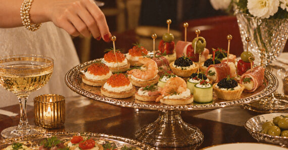 A hand picks an appetizer from a silver tray filled with assorted canapés, including shrimp, caviar, vegetables, and olives, next to a glass of sparkling wine and elegant tableware.