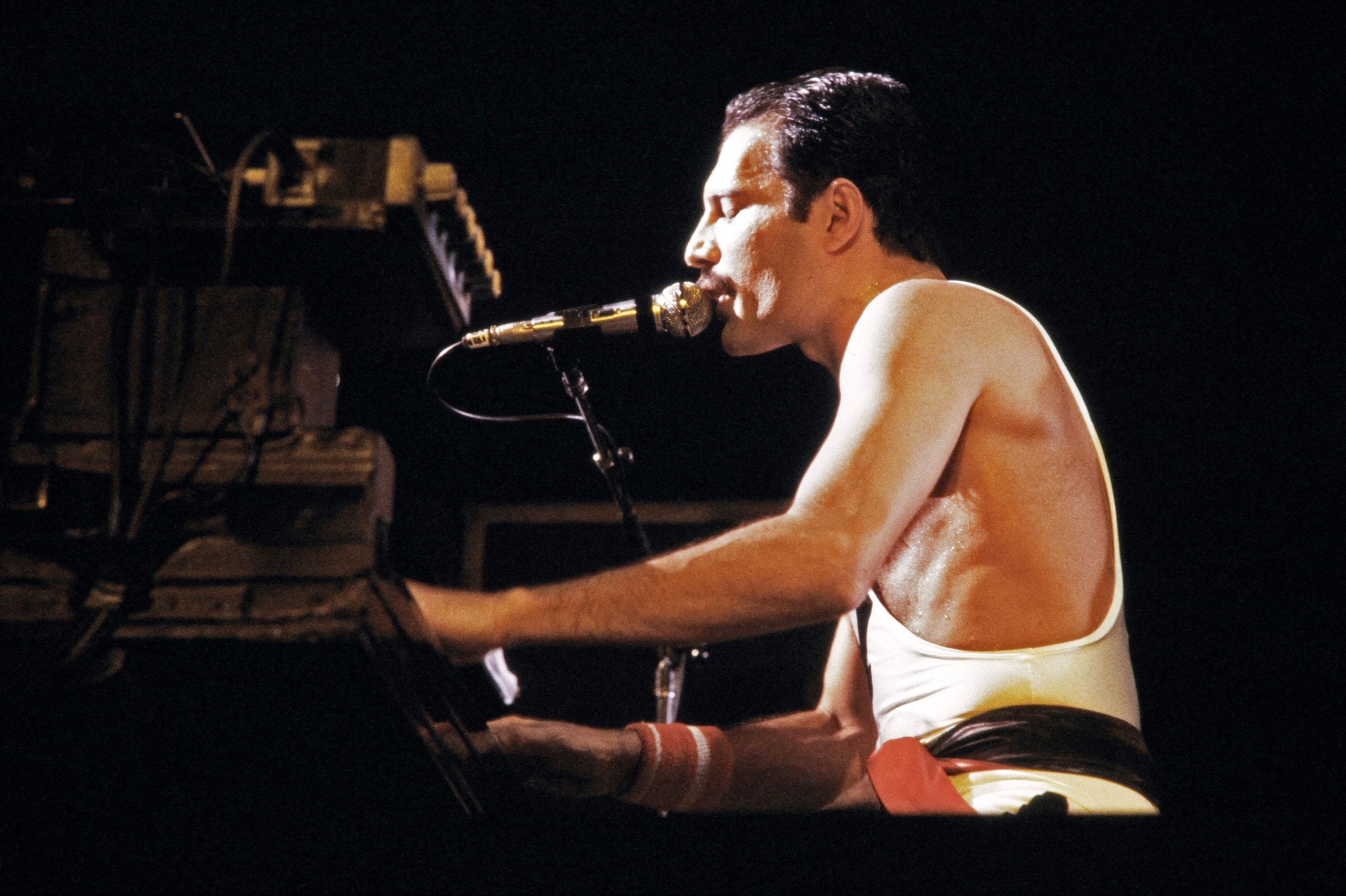 A man in a white tank top sings passionately into a microphone and plays the piano on stage under dramatic lighting.