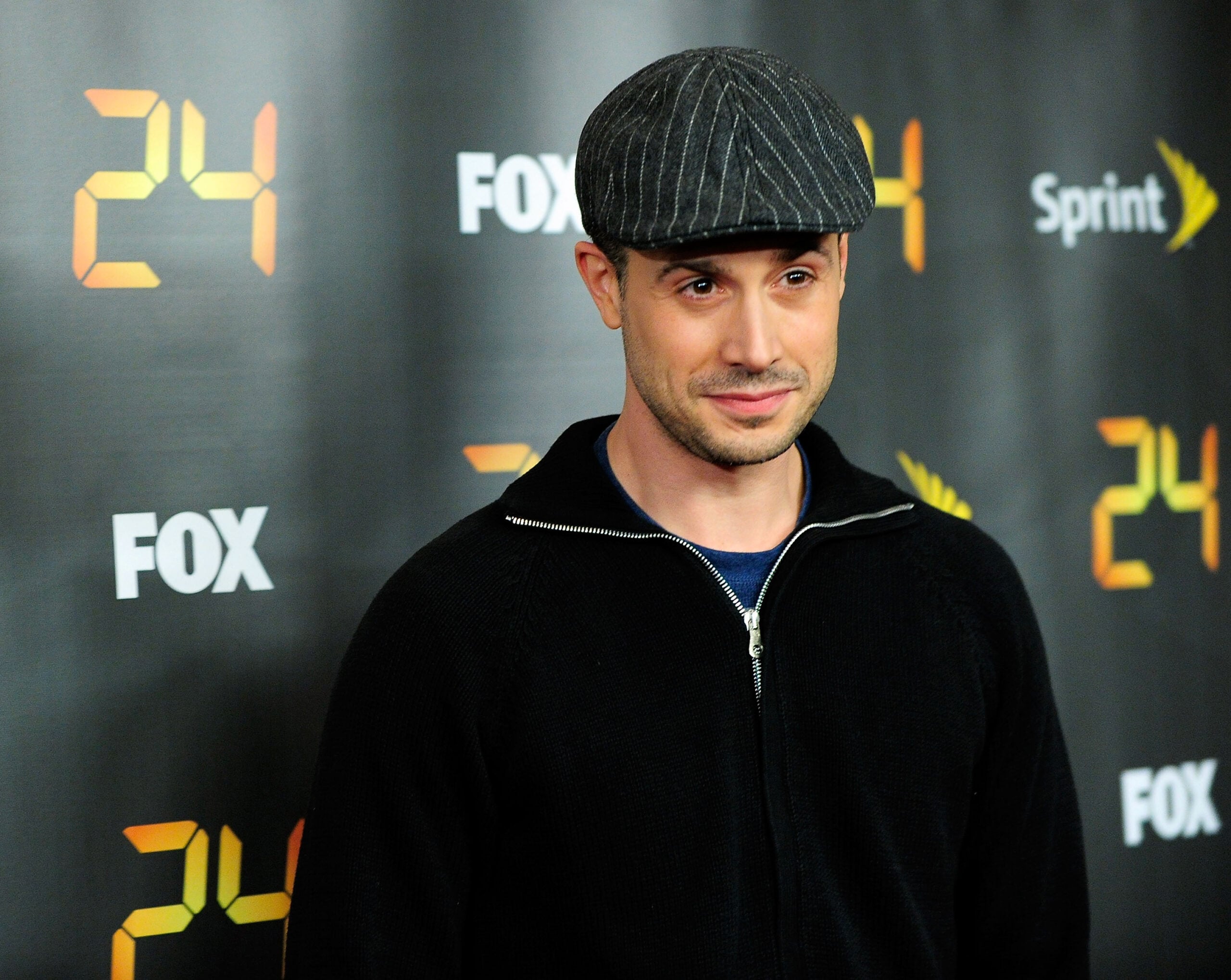 A man in a dark cap and sweater stands in front of a backdrop with “24,” “FOX,” and “Sprint” logos at a media event.