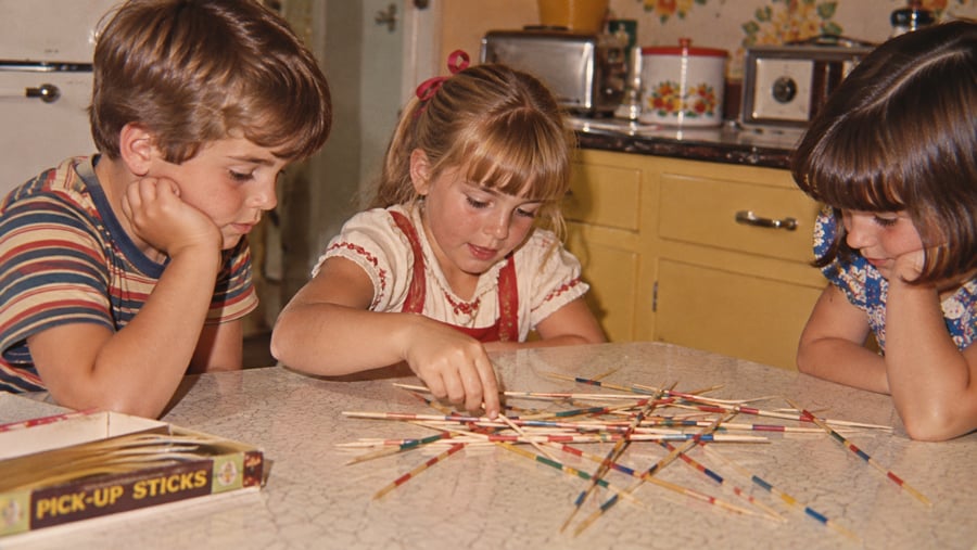 Three children sit at a kitchen table playing Pick-Up Sticks, focused on the game. The vintage kitchen background includes yellow cabinets and a floral container.