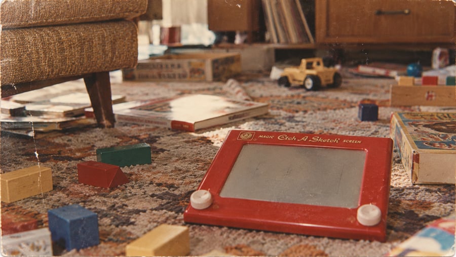 A vintage Etch A Sketch sits on a patterned carpet, surrounded by wooden blocks, board games, and a toy truck. Part of a couch and record player are visible, evoking a nostalgic, cluttered playroom scene.