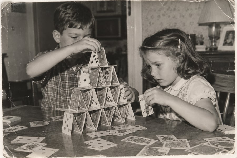 Two children build a pyramid with playing cards on a table, focusing intently. Scattered cards lie around. The image is black and white, evoking a vintage, nostalgic atmosphere.