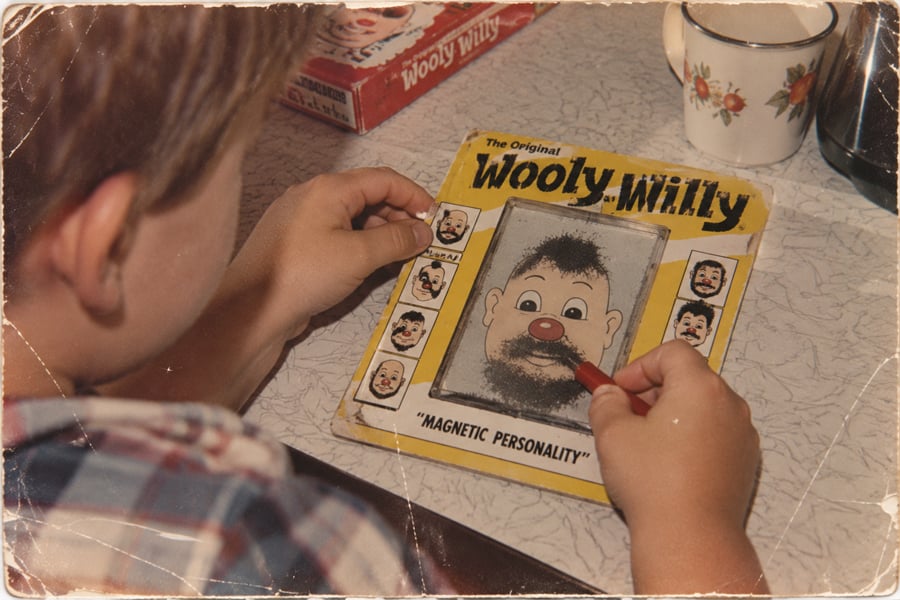 A child uses a red magnetic wand to move black shavings on a Wooly Willy toy, creating facial hair on a cartoon face, at a table with a patterned mug nearby.