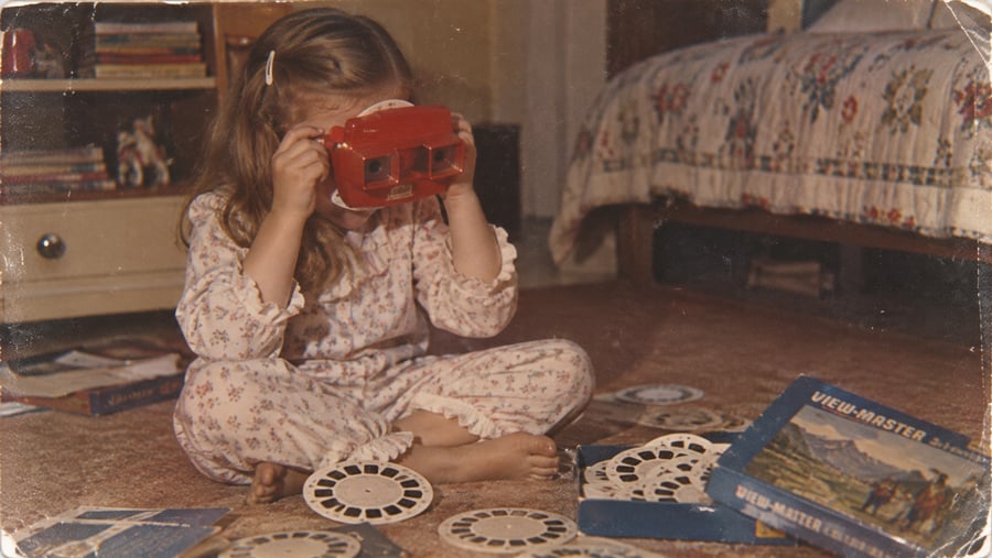 A young girl in pajamas sits cross-legged on the floor, looking through a red View-Master. View-Master reels and boxes are scattered around her. A bed and bookshelf are in the background.
