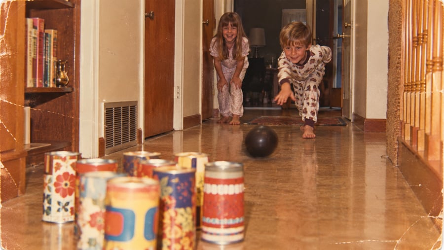 Two children in pajamas play a homemade bowling game in a hallway, using a small black ball and colorful decorated cans as pins. The boy is rolling the ball while the girl smiles behind him.