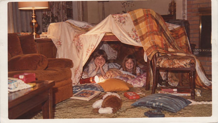 Two children smile inside a homemade blanket fort in a cozy living room, surrounded by pillows, books, and toys, with warm lighting and vintage decor.