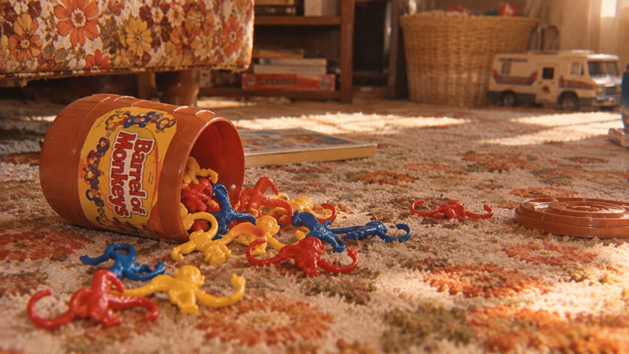 A Barrel of Monkeys toy spills red, blue, and yellow plastic monkeys onto a patterned carpet in a cozy, retro living room with books, a basket, and a toy RV in the background.
