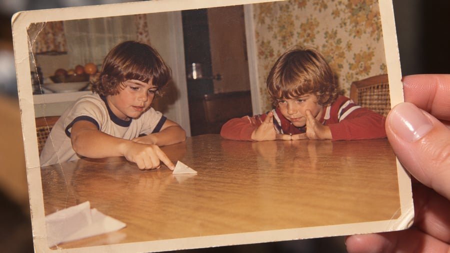 A hand holds an old photo of two children at a wooden table, focused on playing with a paper triangle. The background shows a kitchen with fruit and floral wallpaper.
