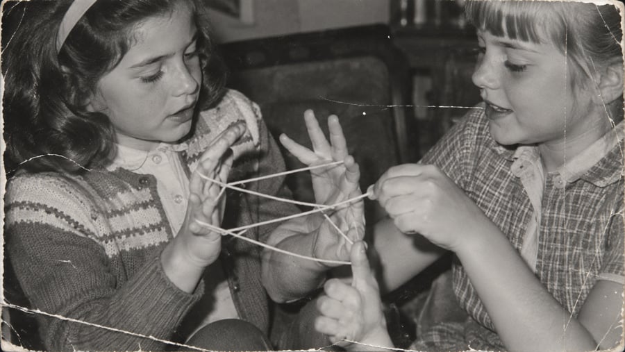 Two young girls play the string game “cat’s cradle,” focusing on the string patterns between their fingers. The black and white photo has a vintage, slightly worn look.