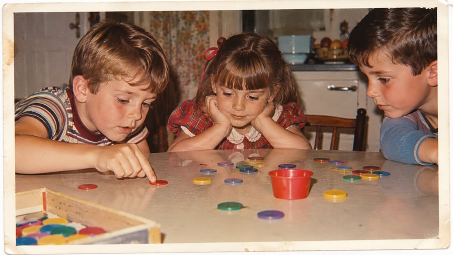 Three children sit around a table, playing with colorful plastic chips. One boy points at a chip while the girl in the center watches closely and another boy looks on. A red cup and a box are also on the table.