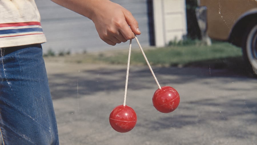 A person holds a pair of red toy balls connected by a white string, known as Clackers, while standing outdoors on a paved surface.