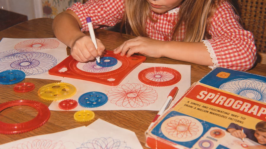 A child in a red checkered dress draws colorful spiral patterns with a Spirograph set on a wooden table, surrounded by gears, pens, and finished designs.