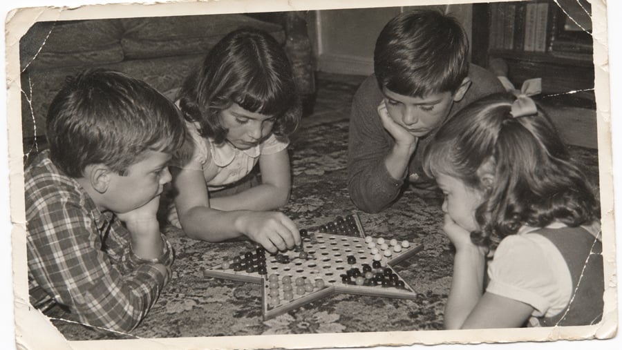 Four children lie on the floor around a star-shaped Chinese checkers board, concentrating on the game. The photo has a sepia tone and worn edges, giving it a nostalgic, vintage feel.