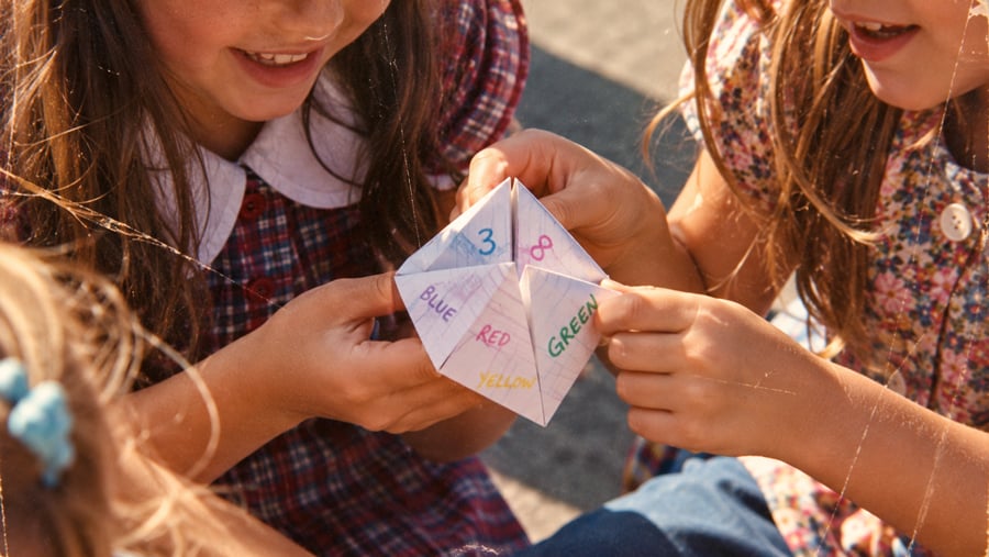 Two children sit closely together, smiling and playing with a colorful paper fortune teller outside on a sunny day. The fortune teller displays numbers and color names written in different colors.
