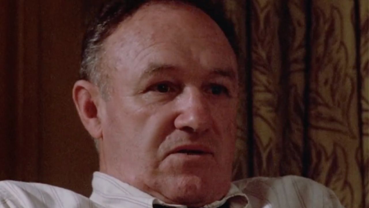 A middle-aged man with short hair sits indoors, wearing a striped shirt. The background shows patterned curtains. He has a neutral expression and is looking slightly off-camera.