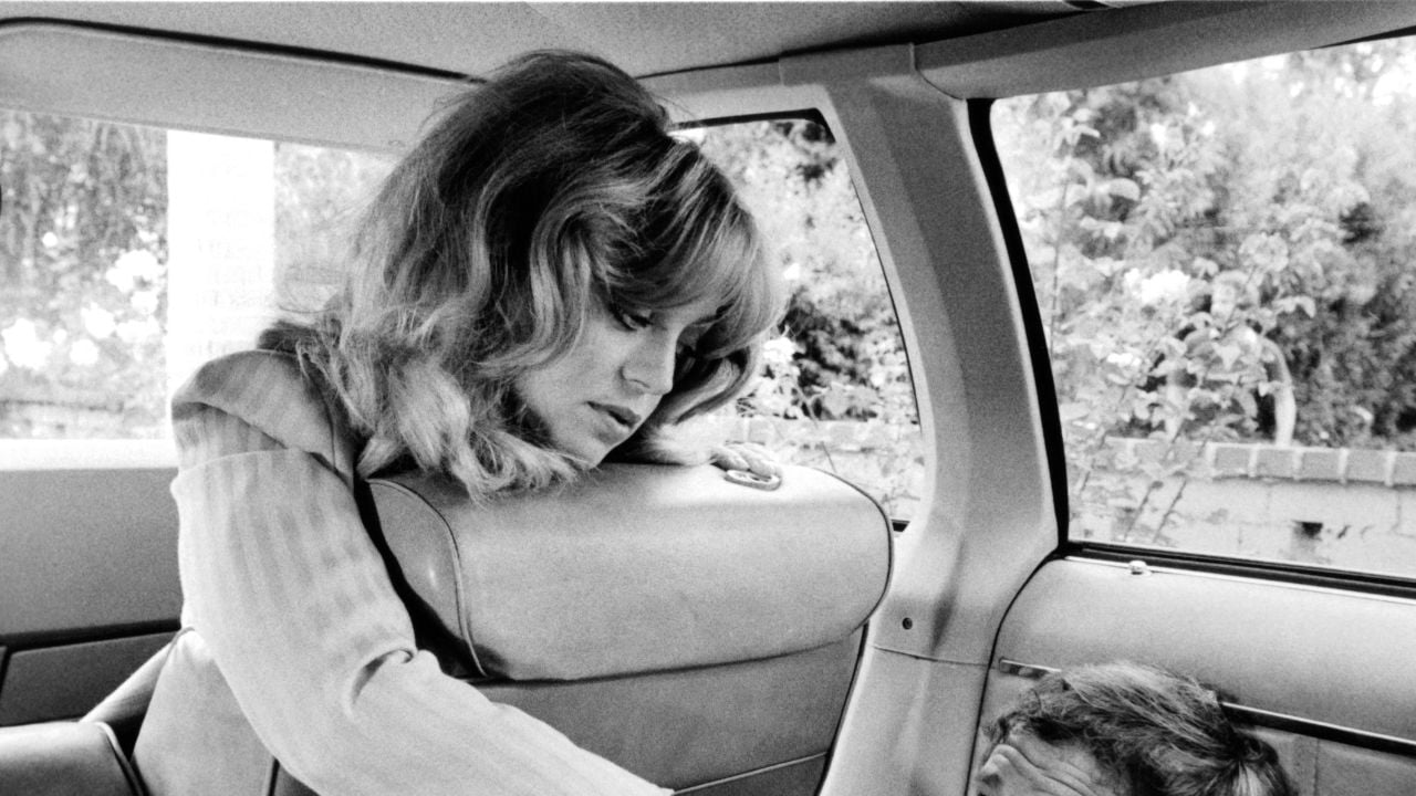 A woman with wavy hair leans over the front seat of a car, looking down at someone partially visible in the backseat. The image is black and white.