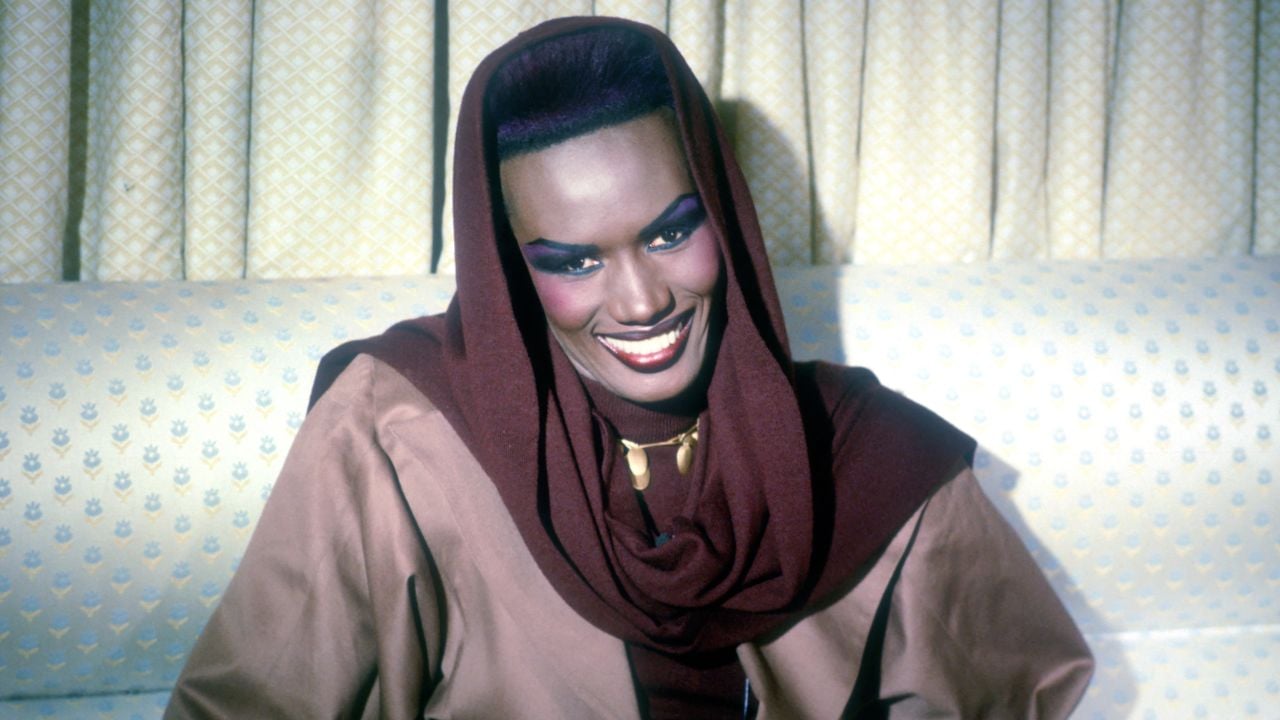 A person with dramatic makeup, wearing a brown and maroon hooded outfit, smiles while sitting on a light patterned sofa with a cream-colored curtain in the background.