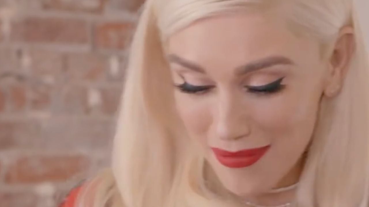 A person with long blonde hair and red lipstick looks downward, smiling softly, with a blurred brick wall in the background.