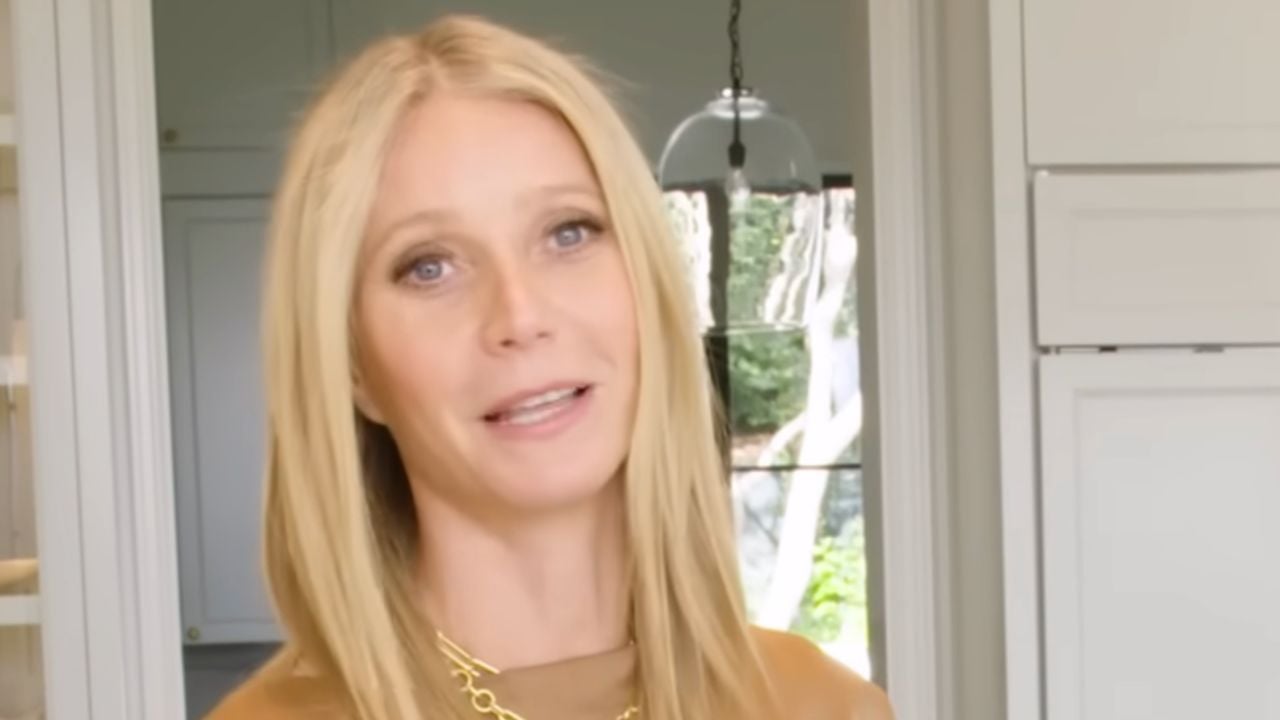 A woman with long straight blonde hair, wearing a brown top and gold necklace, stands indoors in a well-lit modern kitchen with white cabinets and a glass pendant light.