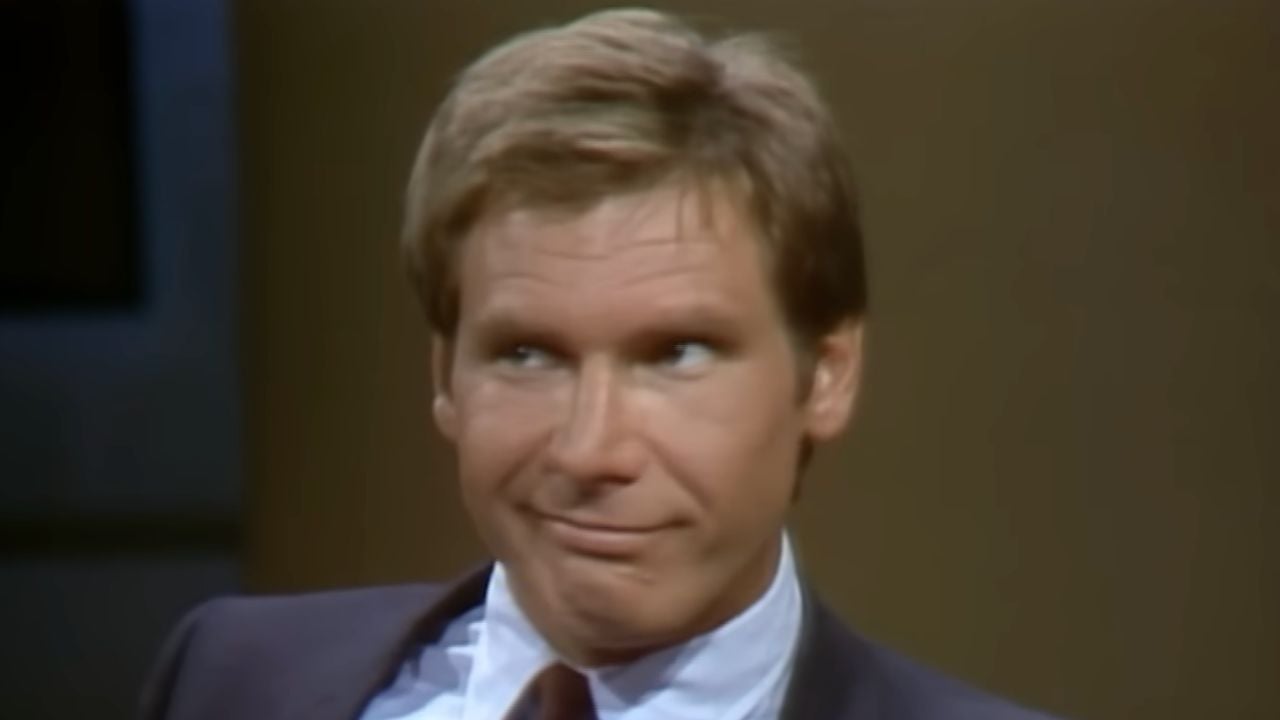 A man in a suit sits indoors, looking to the side with a smirk and raised eyebrow, appearing amused or skeptical.