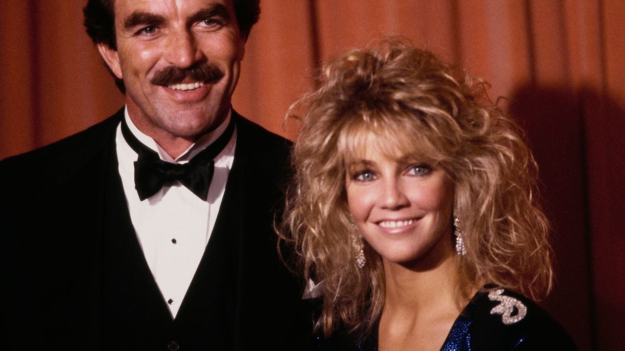 A man in a tuxedo with a mustache stands beside a woman with voluminous blond hair and dangling earrings, both smiling against a reddish curtain backdrop.
