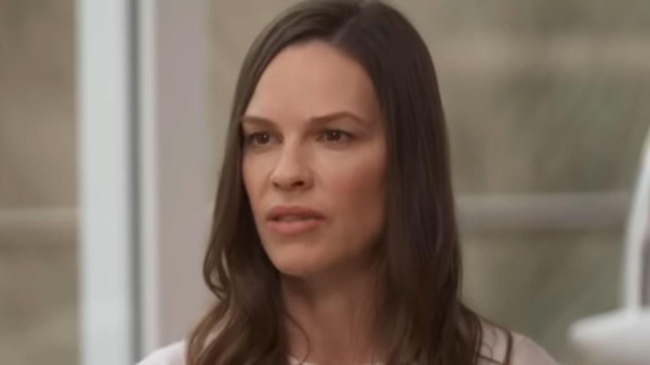 A woman with long brown hair and a serious expression is looking slightly to the side. She is indoors, with a blurred window and neutral background behind her.