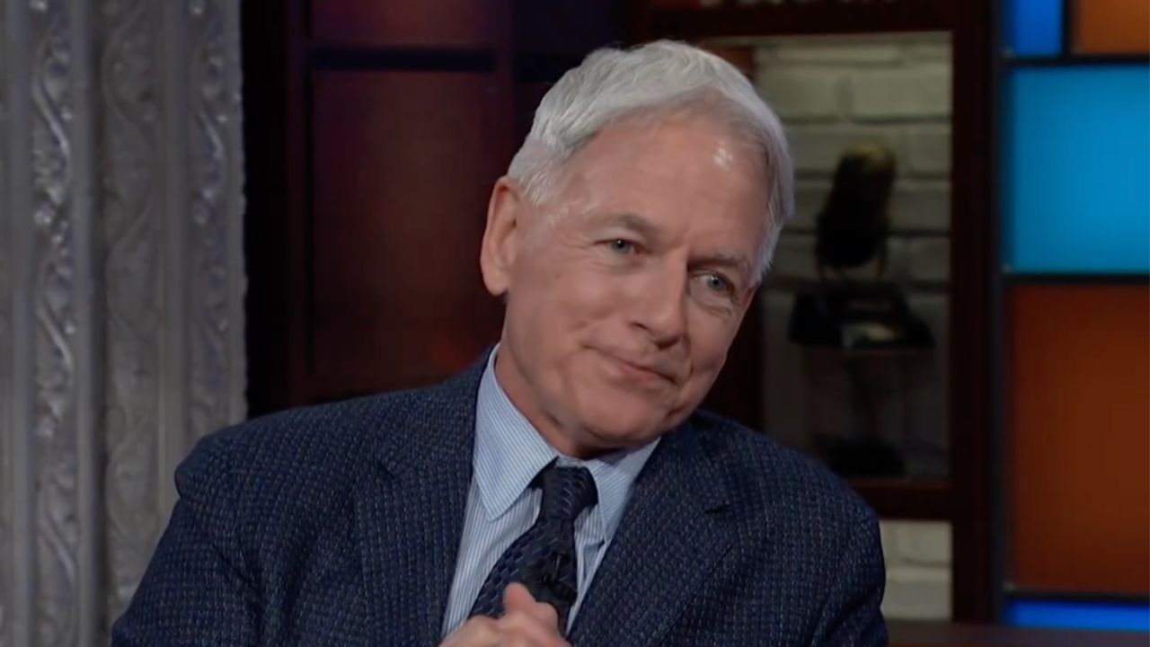 An older man with gray hair, wearing a blue suit and tie, sits indoors with his hands clasped and a slight smile. Colorful panels and a blurred background are visible behind him.