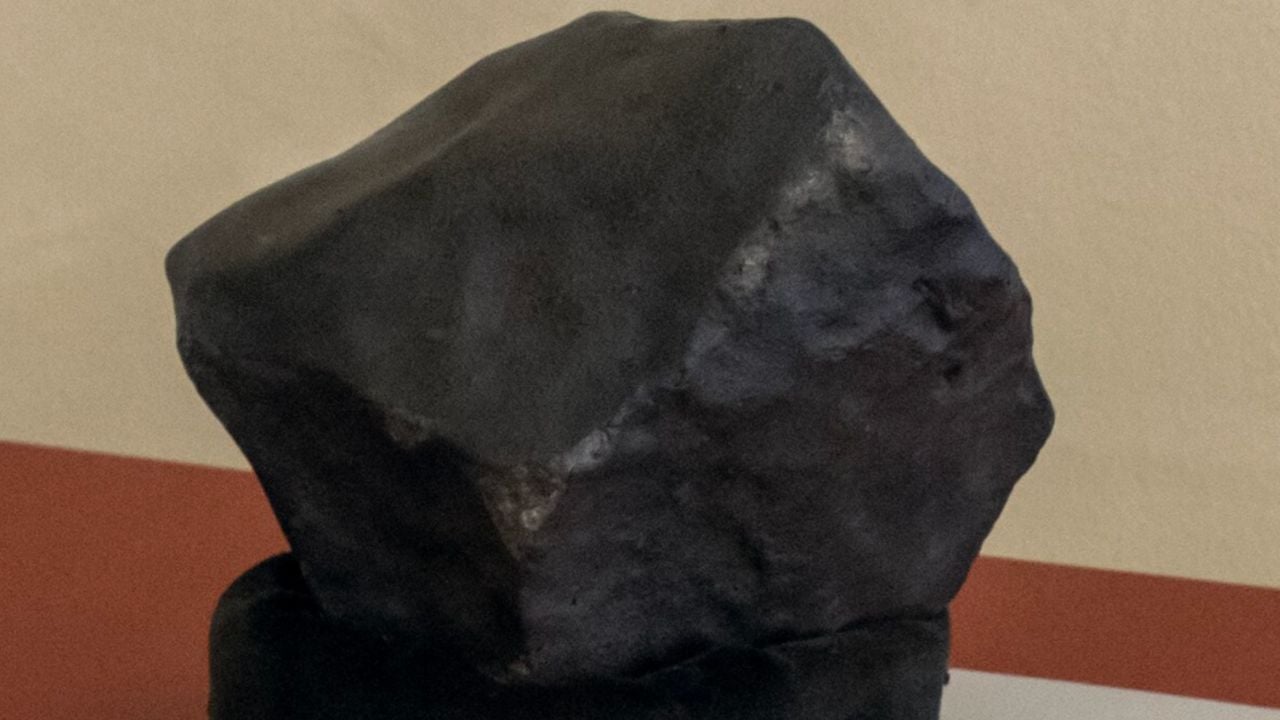 A rough, dark, irregularly shaped rock or mineral specimen is displayed on a stand against a beige and red background.