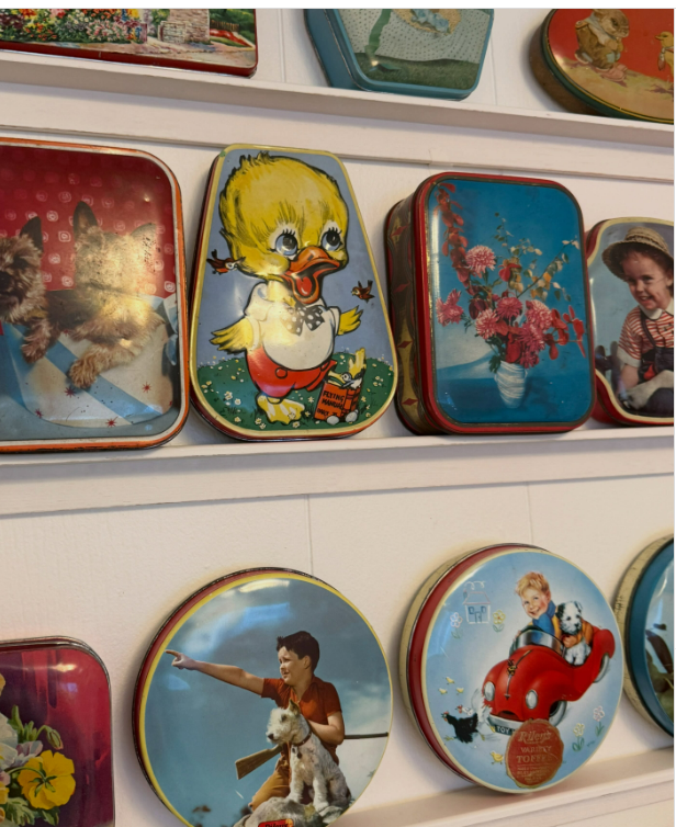 Colorful vintage tins with various illustrations, including animals, flowers, and people, are displayed on white shelves in two rows. The tins are round, rectangular, and unique shapes.