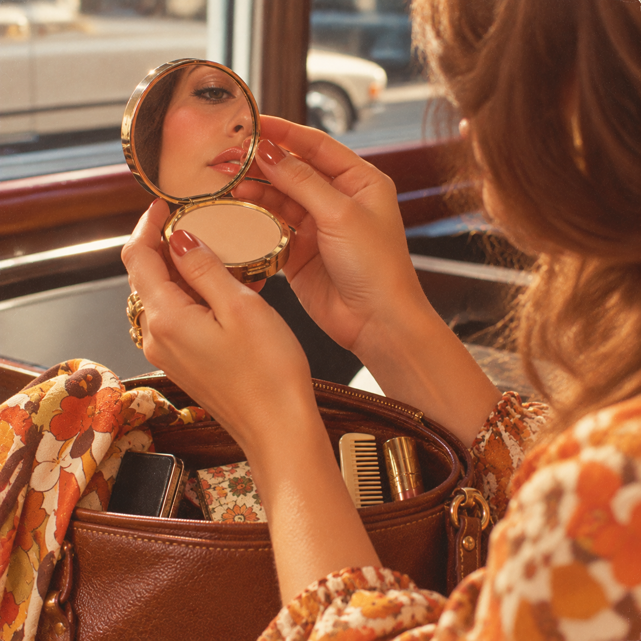 A woman with painted nails holds a compact mirror, looking at her reflection. Her floral blouse and brown handbag, filled with a comb and phone, are visible on her lap in sunlight.