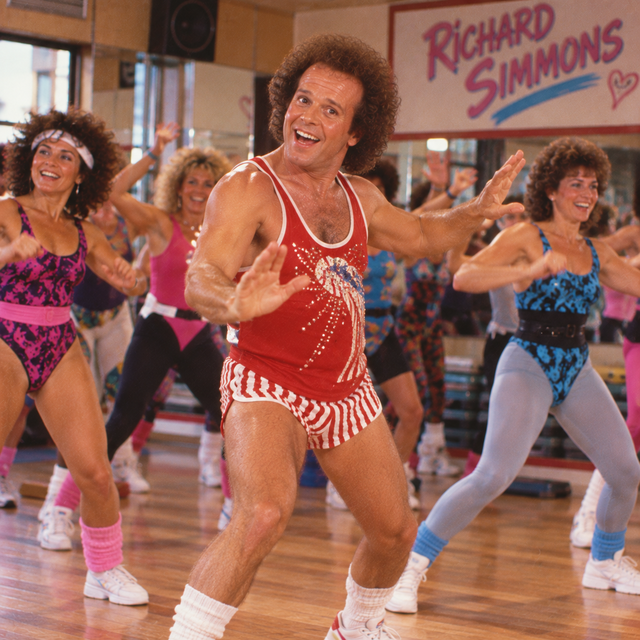 A group of people in colorful workout clothes exercise in a gym, led by a man in a red tank top and striped shorts. A sign reading “Richard Simmons” is visible in the background.