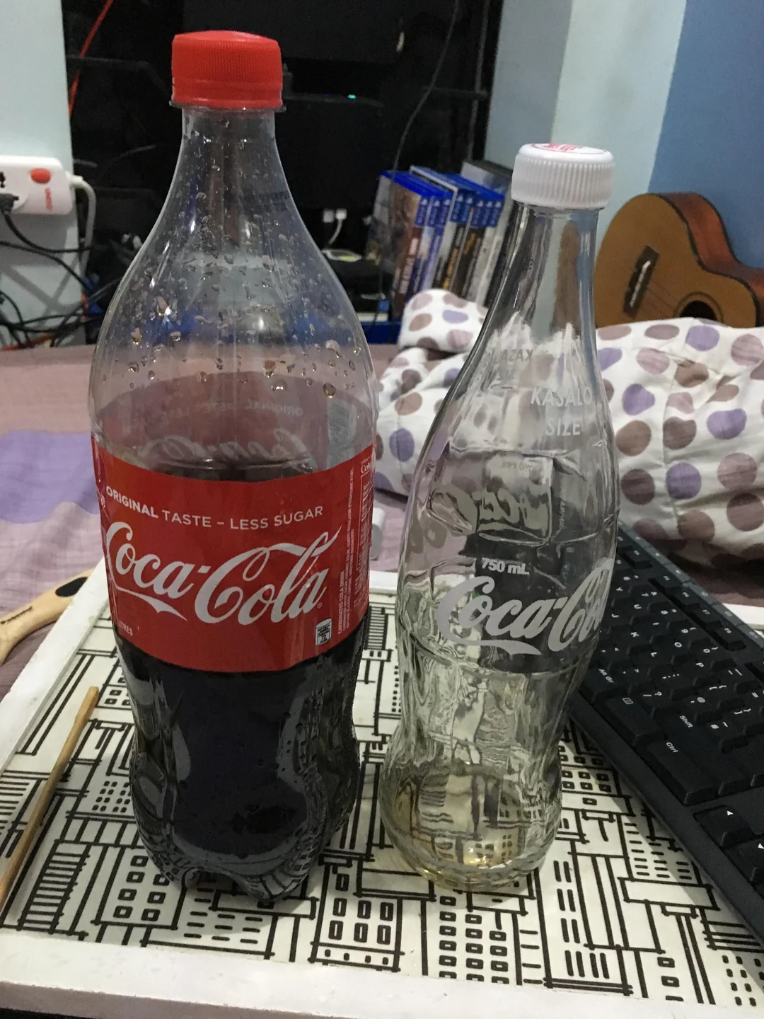 A plastic Coca-Cola bottle with a red cap stands next to an empty glass Coca-Cola bottle with a white cap on a cluttered desk with a keyboard, game cases, and a guitar in the background.