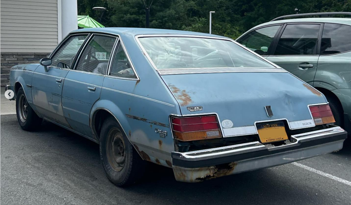 A blue, older-model Buick sedan with visible rust and blemishes is parked in a lot next to a green vehicle. The car has a yellow license plate and some wear on the rear bumper and body.