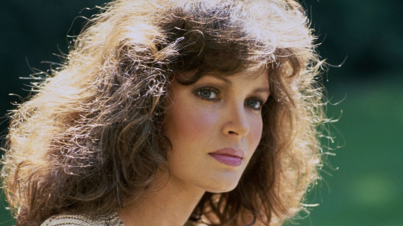 A woman with voluminous brown hair, light makeup, and green eyes looks toward the camera with a neutral expression. The background is blurred and green, suggesting an outdoor setting.