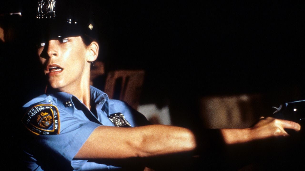 A police officer in uniform, with a tense expression, points a handgun to the right. The background is dark and shadowy.