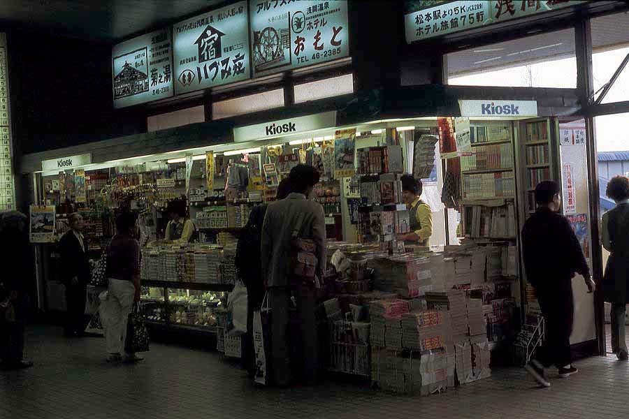 A small kiosk at a train station in Japan sells snacks, drinks, and magazines. Several people browse the items, and signs with Japanese text are displayed above the kiosk.
