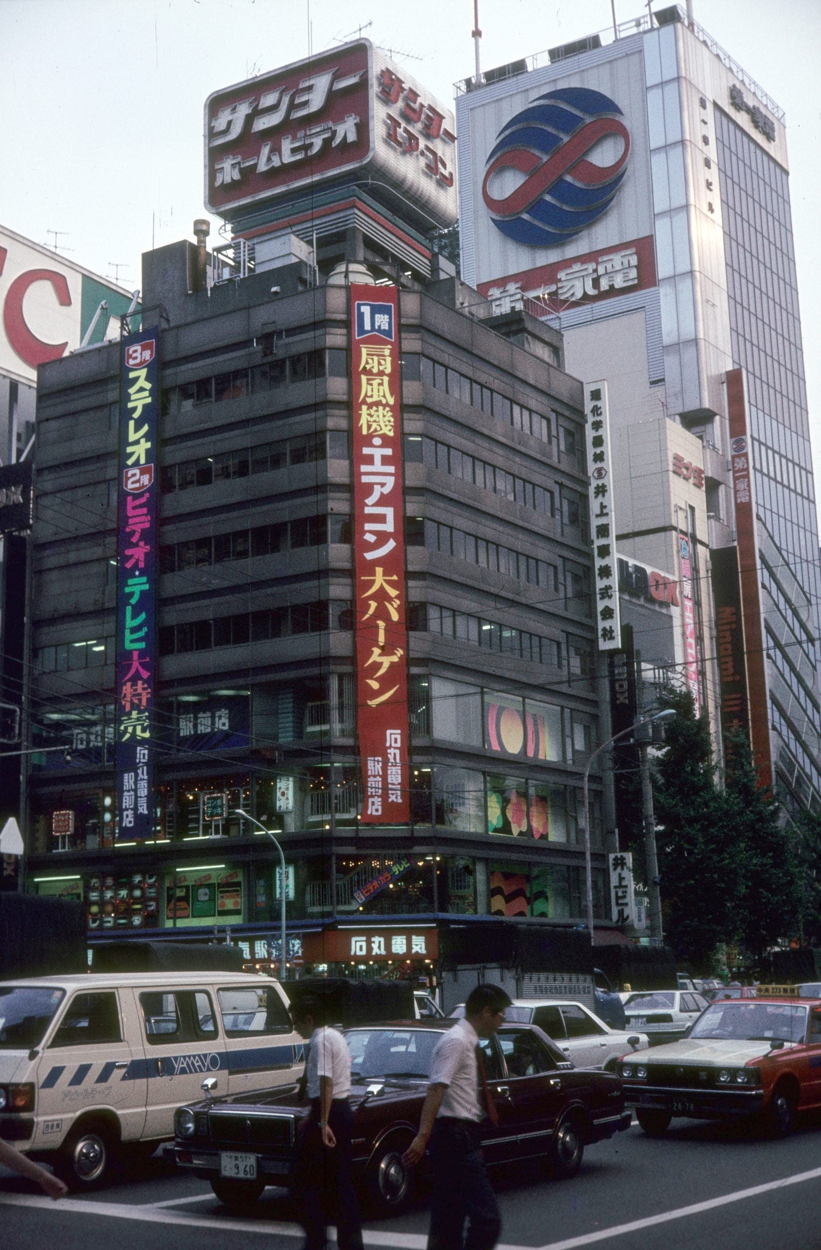 Busy Tokyo street scene with pedestrians crossing, cars stopped, and tall buildings covered in colorful Japanese signs and advertisements.