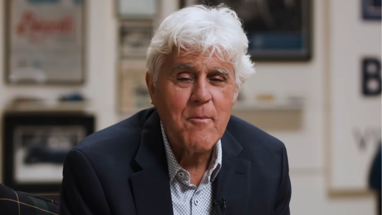 An older man with white hair wearing a dark blazer and light shirt sits indoors, smiling slightly. The background is blurred with framed pictures and posters on the wall.