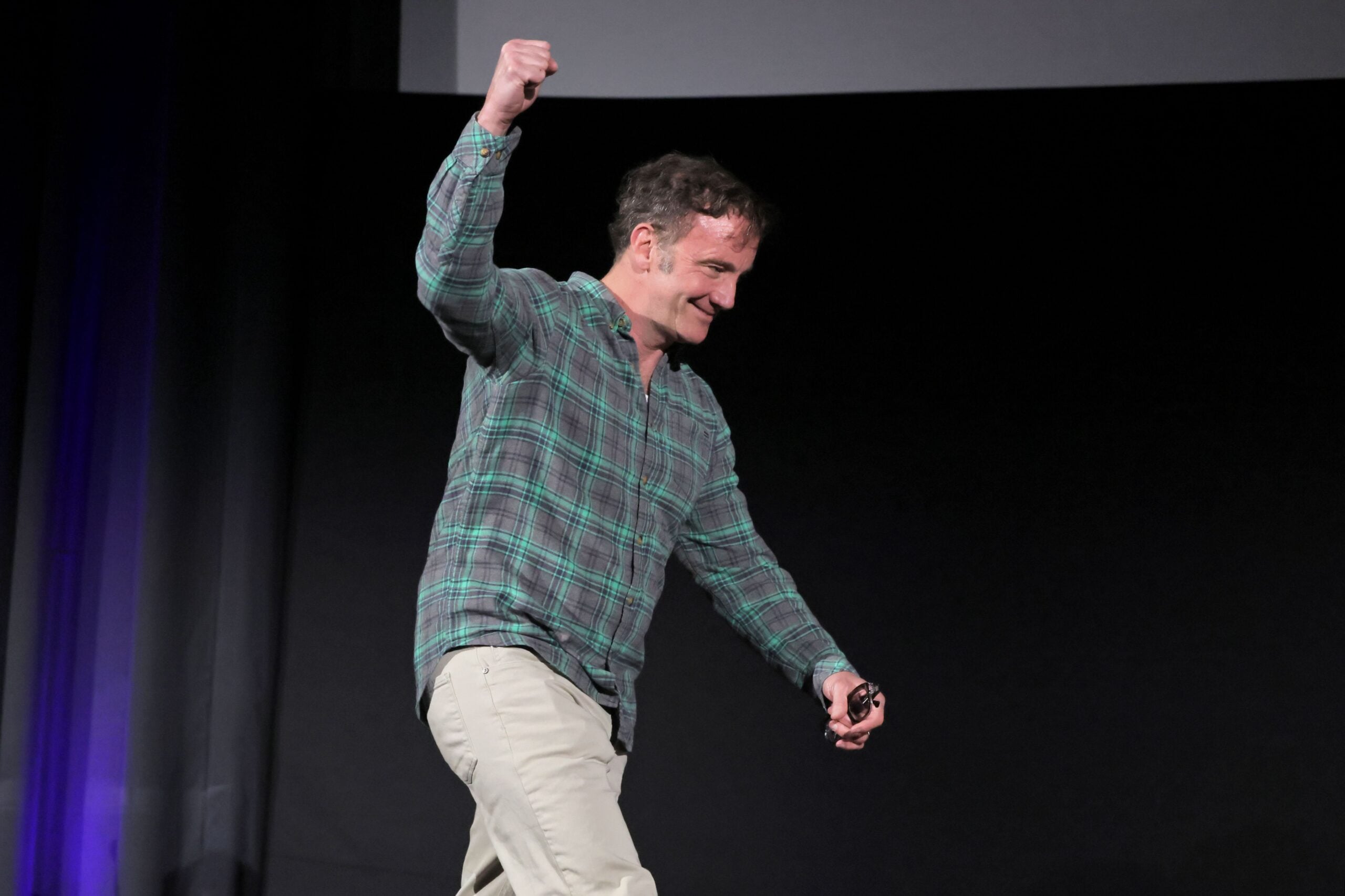 A man in a green plaid shirt and beige pants smiles, raising one fist triumphantly while walking on stage against a dark background.