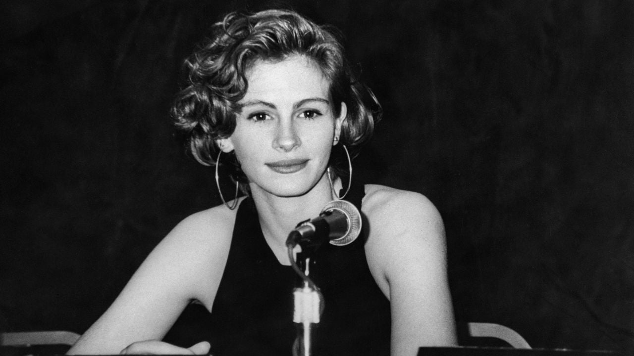 A woman with short, wavy hair sits behind a microphone at a table, wearing a sleeveless top and large hoop earrings, looking directly at the camera against a dark background.