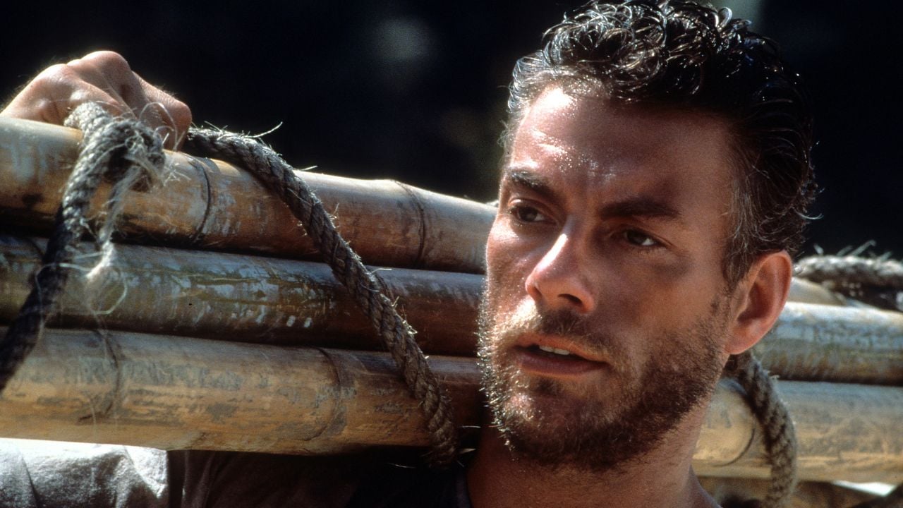 A man with short, wet hair and a beard carries several bamboo poles on his shoulder, looking into the distance with a serious expression.