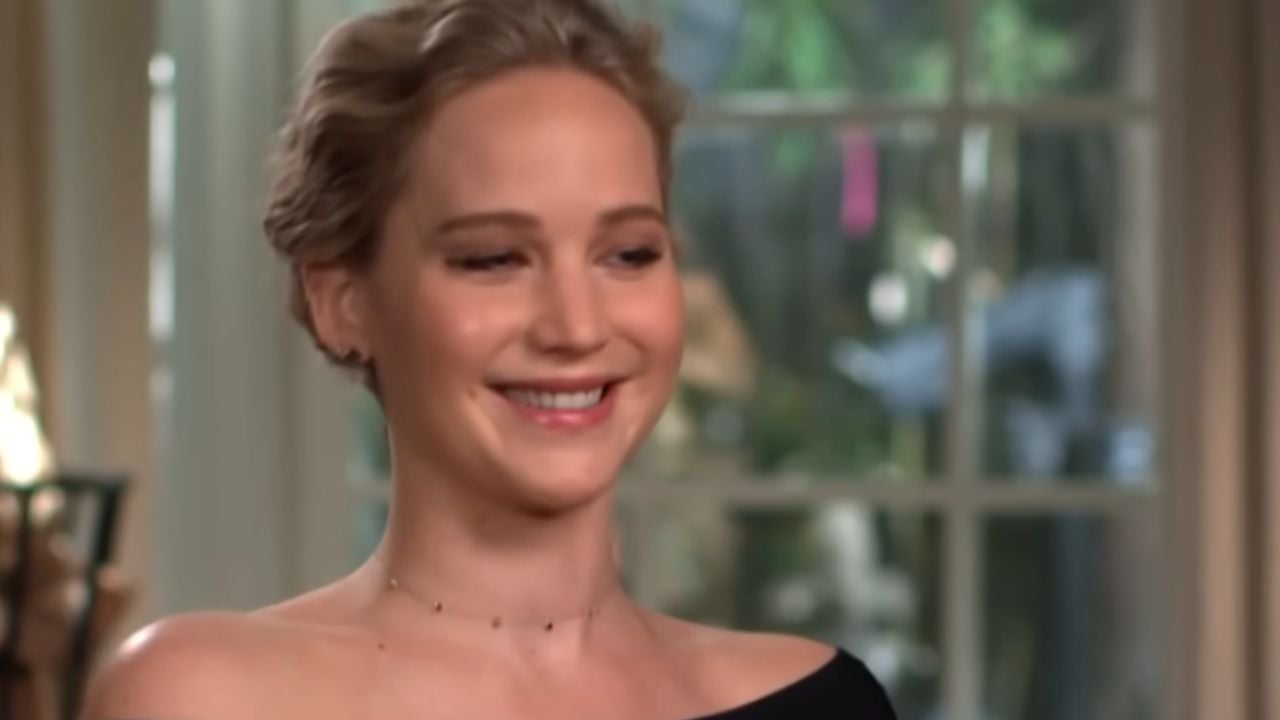 A woman with short blonde hair wearing an off-shoulder black top smiles in a softly lit room with large windows and greenery outside.