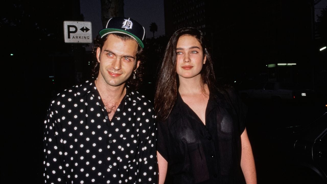 A man in a polka dot shirt and a woman in a black top stand together at night on a city street, with a parking sign and buildings in the background.