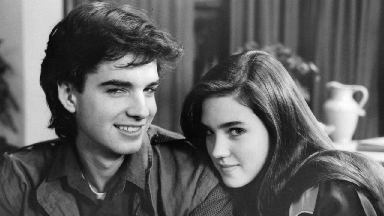 A young man and woman sit close together, smiling and looking into the camera. The image is black and white, with curtains and a jug visible in the background.