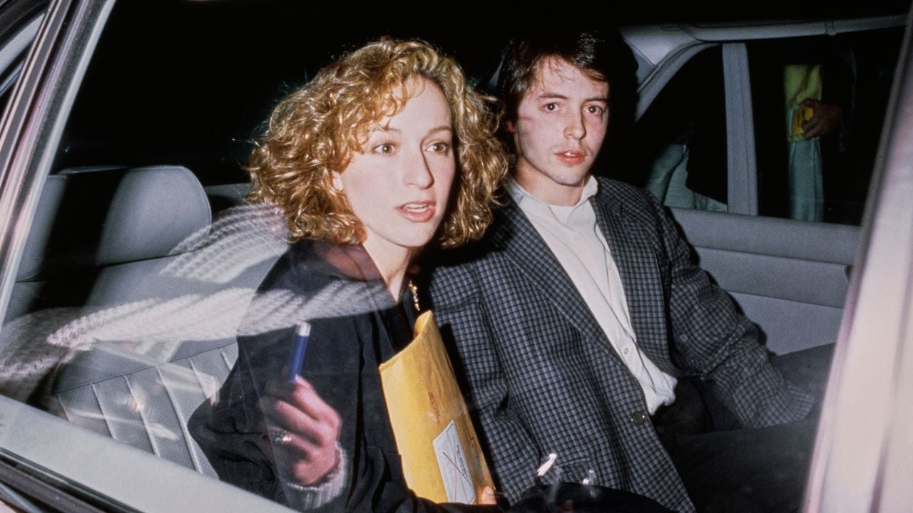 Two people sit in the backseat of a car at night. The woman holds a cigarette and a yellow envelope, while the man sits beside her, looking forward. Both have serious expressions.