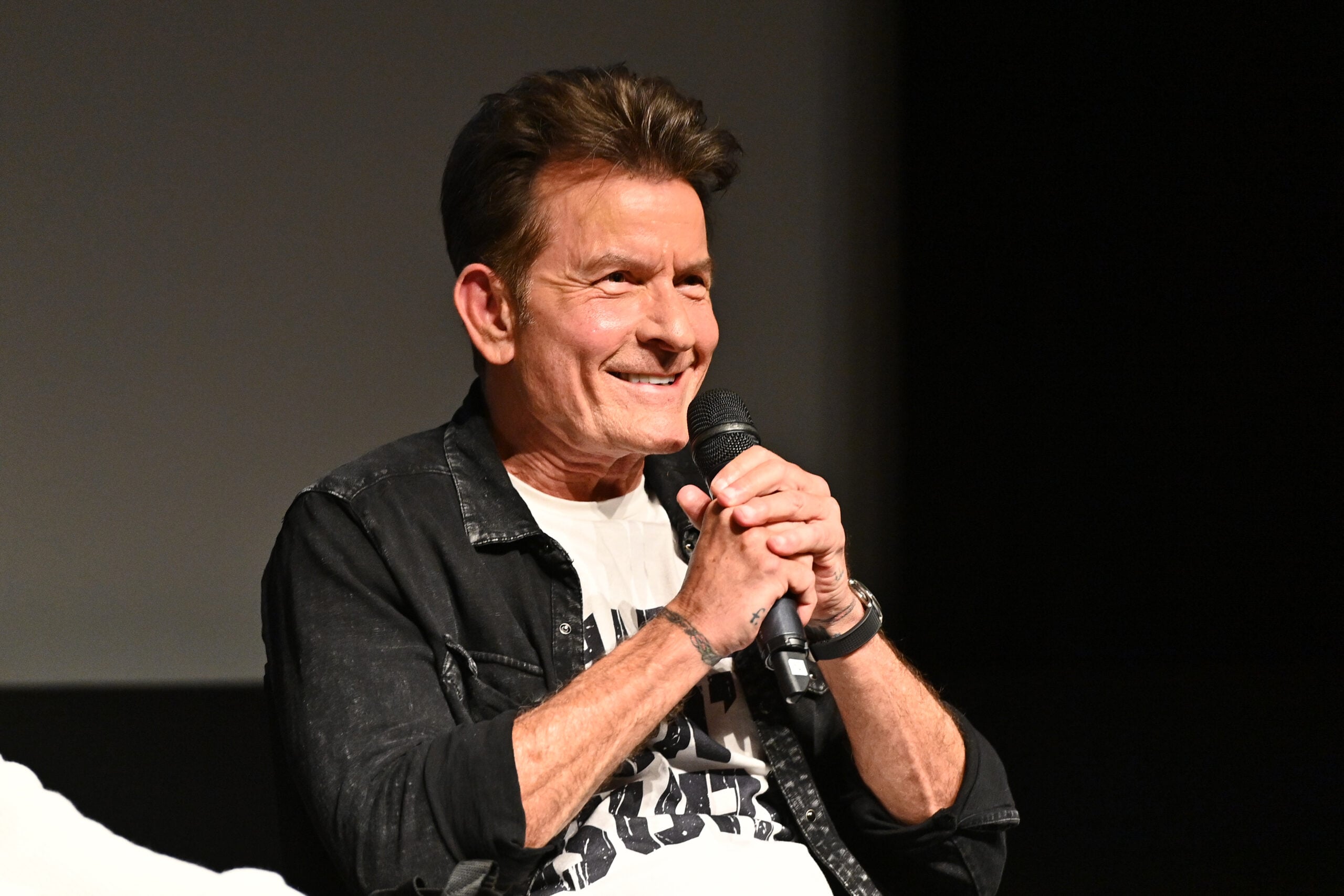 A man with short brown hair smiles while holding a microphone, wearing a black jacket over a graphic t-shirt, seated on a stage with a dark background.