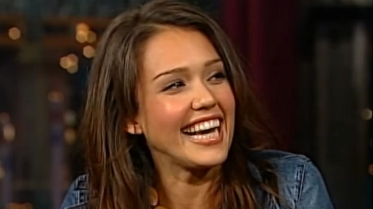 A woman with long brown hair smiles widely, showing her teeth. She is wearing a denim jacket and is looking slightly to her left. The background is blurred with warm lights.