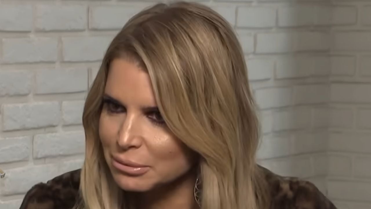 A person with long blonde hair and dramatic makeup is sitting in front of a white brick wall, looking slightly to the side with a neutral expression.