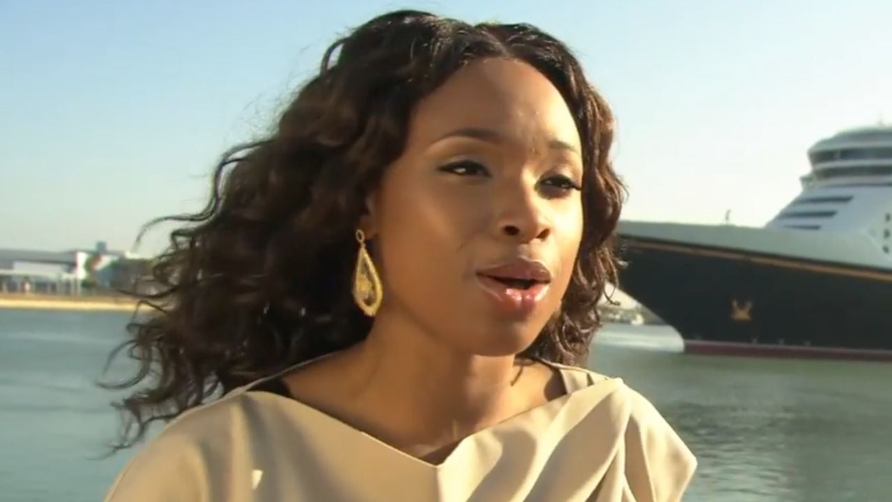 A woman with wavy hair and gold earrings stands outdoors near water, with a large cruise ship and clear sky in the background.