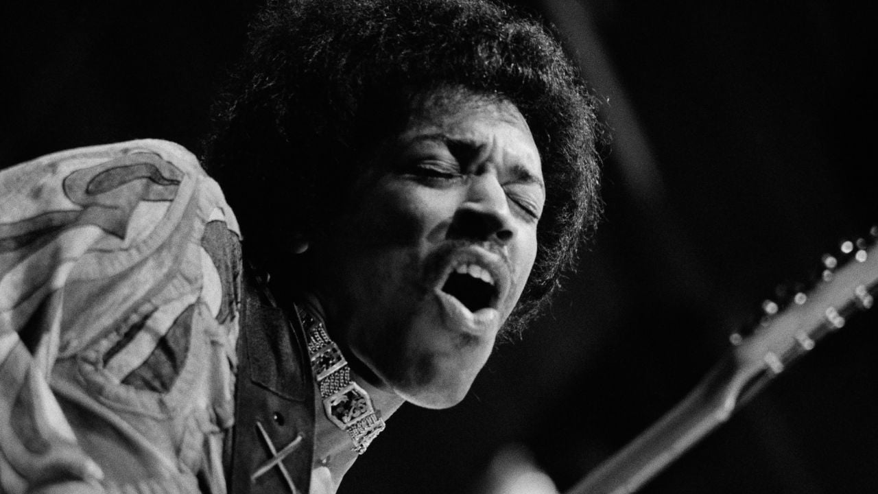 A Black man with an afro hairstyle passionately singing or playing guitar on stage, wearing a patterned shirt and a decorative scarf, shown in dramatic black and white lighting.